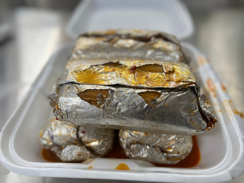 May include: A stack of tamales wrapped in silver foil, presented in a white foam container. The tamales have a golden-brown filling visible through the foil. The container has a small amount of sauce at the bottom.