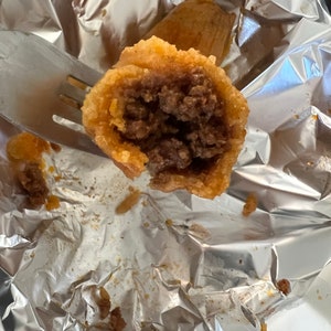 May include: A close-up of a cut-open, golden-brown fried food item revealing a dark brown meat filling. The food is held by a fork, and rests on a crumpled aluminum foil surface. Another similar item is visible in the background.