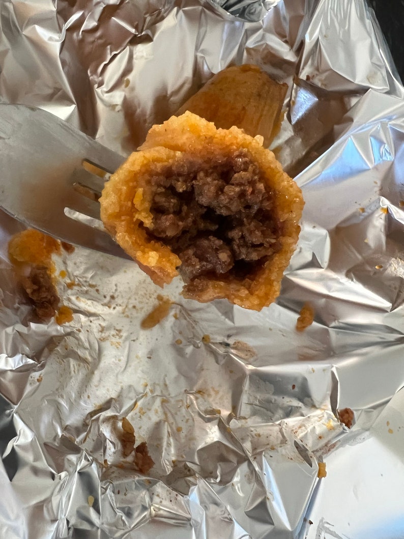 May include: A close-up of a cut-open, golden-brown fried food item revealing a dark brown meat filling. The food is held by a fork, and rests on a crumpled aluminum foil surface. Another similar item is visible in the background.