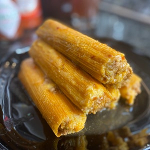 May include: A close-up of a stack of golden-brown tamales on a black plate. The tamales are wrapped in corn husks and filled with a savory meat filling. The dish is a traditional Mexican food.