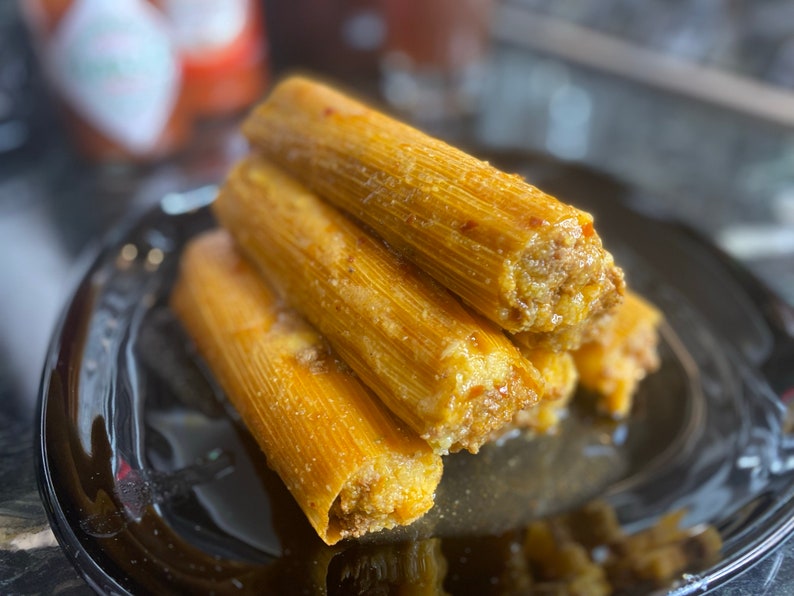 May include: A close-up of a stack of golden-brown tamales on a black plate. The tamales are wrapped in corn husks and filled with a savory meat filling. The dish is a traditional Mexican food.
