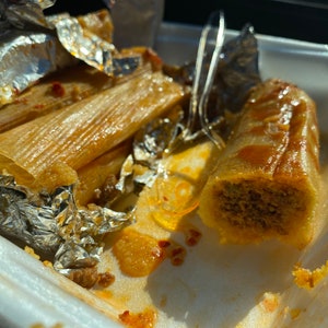 May include: A close-up shot of tamales in a white foam container. The tamales are wrapped in corn husks and partially covered in aluminum foil. One tamale is cut open, revealing the filling. A fork rests in the container.