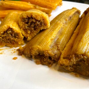 May include: A plate of cooked tamales, a traditional Mexican dish. The tamales are wrapped in corn husks and filled with a savory meat filling. The husks are a light tan color, and the filling is a darker brown. Some tamales are cut open, revealing the filling.