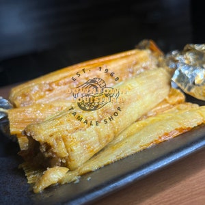 May include: Close-up of several golden-brown tamales on a black rectangular plate. The tamales are wrapped in corn husks and appear to be filled with meat. The image includes the text "Downtown Louisiana Tamale Shop Est. 1968".