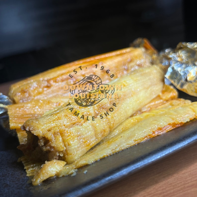May include: Close-up of several golden-brown tamales on a black rectangular plate. The tamales are wrapped in corn husks and appear to be filled with meat. The image includes the text "Downtown Louisiana Tamale Shop Est. 1968".