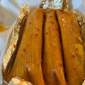 May include: Three golden-brown tamales, wrapped in corn husks, are presented in a white foam container. The tamales are glistening, with visible red specks, and are partially encased in aluminum foil.