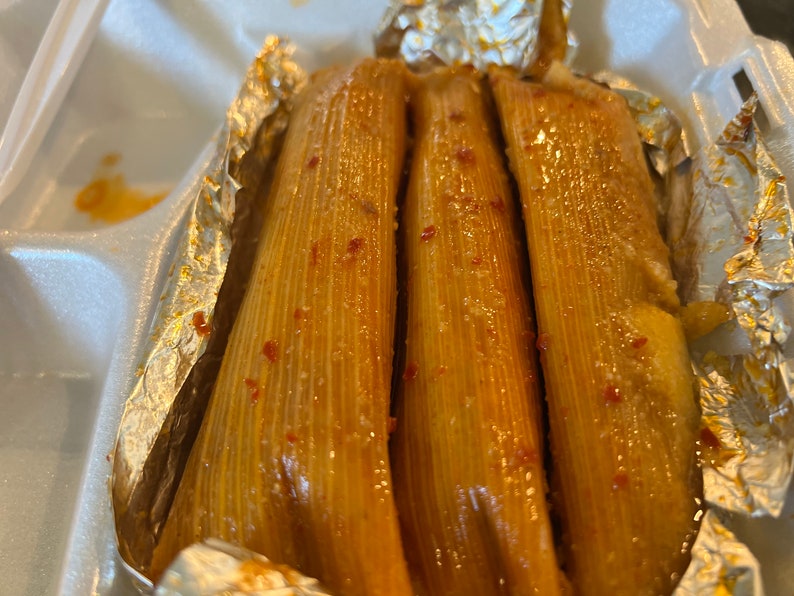 May include: Three golden-brown tamales, wrapped in corn husks, are presented in a white foam container. The tamales are glistening, with visible red specks, and are partially encased in aluminum foil.