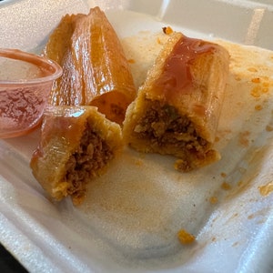May include: A close-up of tamales, a traditional dish, served in a white foam container. The tamales are partially unwrapped, revealing a savory meat filling. A small dish of sauce is visible.
