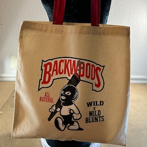 May include: A beige tote bag with red handles features a Backwoods logo with a cartoon character holding a cigar. The text on the bag reads "All Natural" and "Wild 'n Mild Blunts".