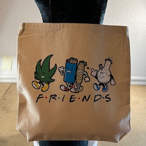 May include: A beige canvas tote bag with a graphic print of four cartoon characters. The characters are a marijuana leaf, a lighter, a joint, and a glass pipe. The text "FRIENDS" is printed below the characters.