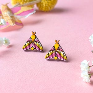 May include: A pair of wooden moth-shaped stud earrings. The earrings feature a yellow head and wing accents, with pink and black details. The earrings are set against a pink background, with decorative flowers.