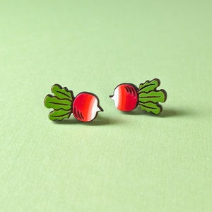 Peut inclure: Une paire de boucles d'oreilles en forme de radis. Chaque boucle d'oreille représente un radis rouge et blanc avec des feuilles vertes. Les boucles d'oreilles sont posées sur un fond vert clair. Elles sont petites et semblent être en bois.