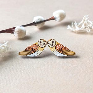 May include: Two wooden owl-shaped earrings. The owls face each other, with a heart-shaped design on their heads. The earrings have a yellow, brown, and white color scheme. A sprig of pussy willow and white decorative elements are in the background.