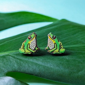 May include: A pair of green frog earrings on a green leaf background. The frogs are facing each other and have white eyes.