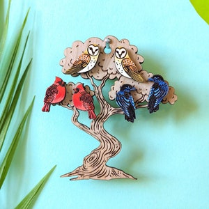 May include: A set of six wooden earrings featuring different bird designs. The earrings are displayed on a wooden tree branch cutout. The birds include a cardinal, a barn owl, and a blue jay.
