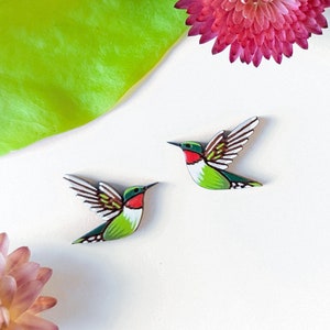 May include: Two wooden hummingbird ornaments with green, white, and red paint. The hummingbirds are in flight with their wings spread.
