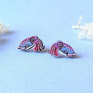 May include: A pair of stud earrings shaped like squids, featuring a gradient of purple and pink colors with white spots on the mantle and detailed tentacles. The design is flat and colorful, showcasing a stylized marine creature aesthetic.