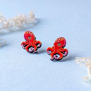 May include: A pair of stud earrings shaped like small octopuses. Each octopus is colored in vibrant shades of red and orange with black outlines and white dots along the tentacles. The design features a flat, detailed surface with a clear, defined shape.