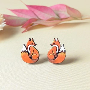 May include: A pair of orange fox stud earrings. Each earring features a stylized fox with a white-tipped tail and black outline. The earrings are set against a cream-colored background with pink and white floral accents.