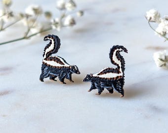 May include: A pair of skunk-shaped earrings. Each earring features a black body with a white stripe down the back and a fluffy tail. The skunks are set against a white background with delicate white flowers.