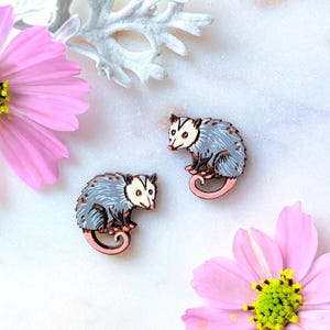 May include: Two opossum-shaped pins with gray fur, pink tails, and white faces. The pins are on a white surface with pink cosmos flowers and silver foliage. The pins are likely made of wood or a similar material.