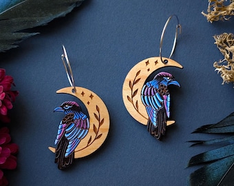 May include: A pair of dangle earrings featuring wooden crescent moon charms with laser-engraved leaf and star details. Each moon holds a perched raven with iridescent blue and purple plumage. The charms hang from thin silver-toned metal hoops.
