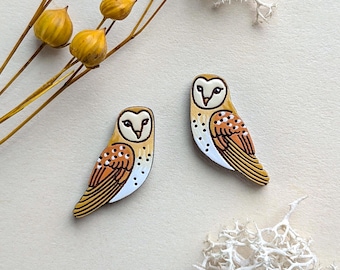 May include: Two barn owl pins featuring a cream-colored face, brown and orange wings with white spots, and a dark brown beak. The owls are depicted in a side profile view with detailed line work, showcasing a smooth finish and distinct avian features.