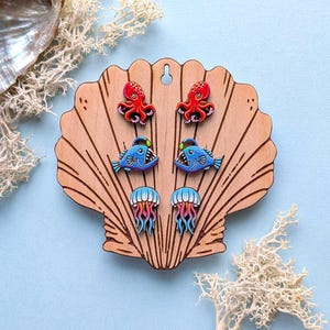 May include: Three pairs of colorful stud earrings displayed on a light wood shell-shaped holder. The top pair features red octopuses, the middle pair shows blue anglerfish with green lures, and the bottom pair depicts blue and pink jellyfish.
