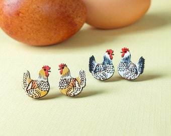 May include: Wooden chicken earrings with red combs and detailed feather patterns. Two brown eggs are in the background, with a light yellow surface. The earrings are in the shape of a hen, with a yellow, white, and black design.