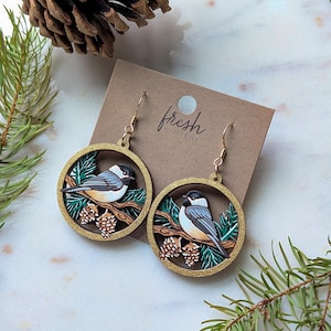 May include: A pair of circular dangle earrings featuring a black-capped chickadee perched on a pine branch with cones. The earrings are mounted on a brown card labeled fresh CUTTERY and have gold-toned metal hooks for pierced ears.