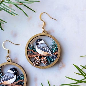 May include: A pair of circular dangle earrings featuring a black, white, and gray chickadee bird perched on a brown branch with pinecones and green pine needles, all encased in a gold-toned frame with gold-toned metal earring hooks.