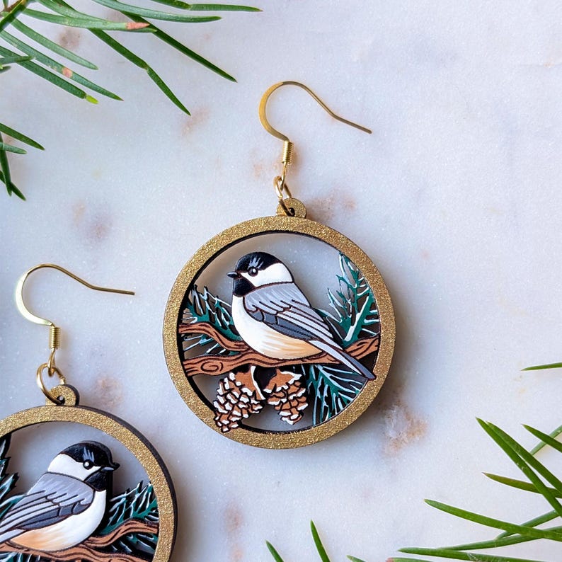May include: A pair of circular dangle earrings featuring a black, white, and gray chickadee bird perched on a brown branch with pinecones and green pine needles, all encased in a gold-toned frame with gold-toned metal earring hooks.