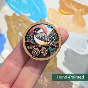 May include: A circular wooden ornament featuring a hand-painted black-capped chickadee perched on a branch with pinecones and needles. The frame is gold-toned with a small loop at the top for hanging. The text Hand-Painted is visible.