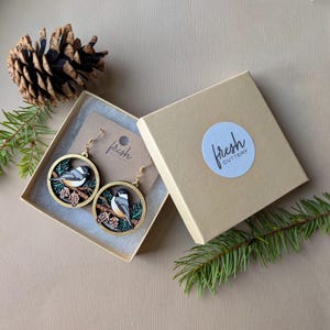 May include: A pair of circular wooden earrings featuring a black-capped chickadee bird perched on a branch with pinecones. The earrings are presented in a tan cardboard gift box with a round sticker labeled fresh cutlery.