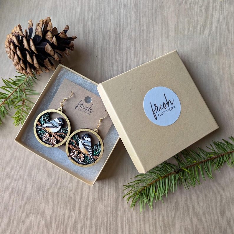 May include: A pair of circular wooden earrings featuring a black-capped chickadee bird perched on a branch with pinecones. The earrings are presented in a tan cardboard gift box with a round sticker labeled fresh cutlery.