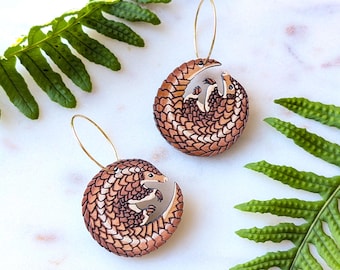 May include: A pair of circular dangle earrings featuring a detailed illustration of a curled-up pangolin with brown scales. Each earring is suspended from a thin gold-toned metal hoop wire, showcasing the animal in a protective, rounded posture.