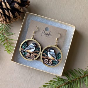 May include: A pair of round dangle earrings featuring a black, white, and tan chickadee bird perched on a branch with pinecones. The earrings are set in a gold-toned circular frame and attached to a card labeled fresh cutlery.