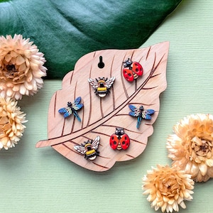 May include: A light brown wooden display board shaped like a leaf, featuring five insect-shaped pins: two red and black ladybugs, two iridescent blue dragonflies, and one yellow and black bumblebee, all arranged on the surface of the wood.