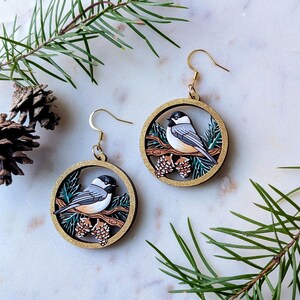 May include: A pair of circular dangle earrings featuring a black-capped chickadee perched on a brown branch with pine cones and green needles. The round frames have a gold glitter finish and gold-toned metal ear wires for pierced ears.