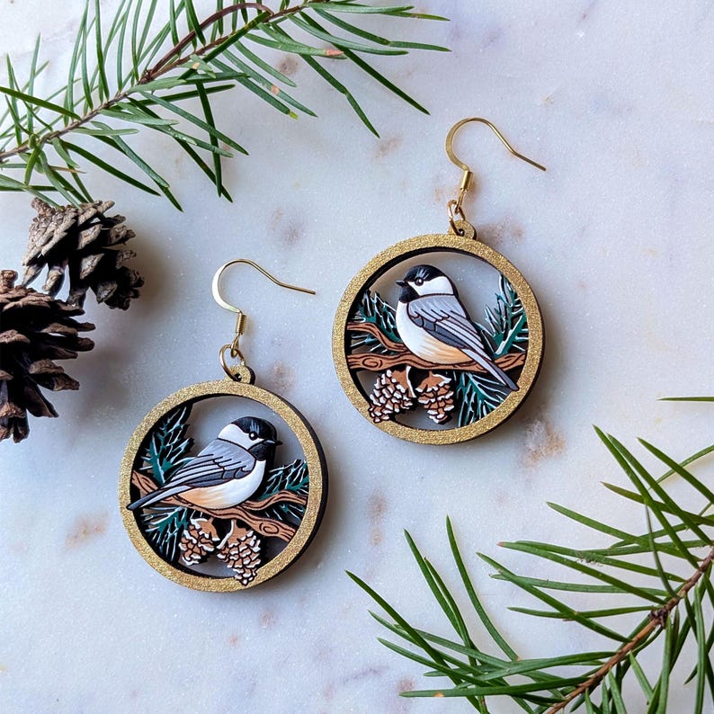 May include: A pair of circular dangle earrings featuring a black-capped chickadee perched on a brown branch with pine cones and green needles. The round frames have a gold glitter finish and gold-toned metal ear wires for pierced ears.