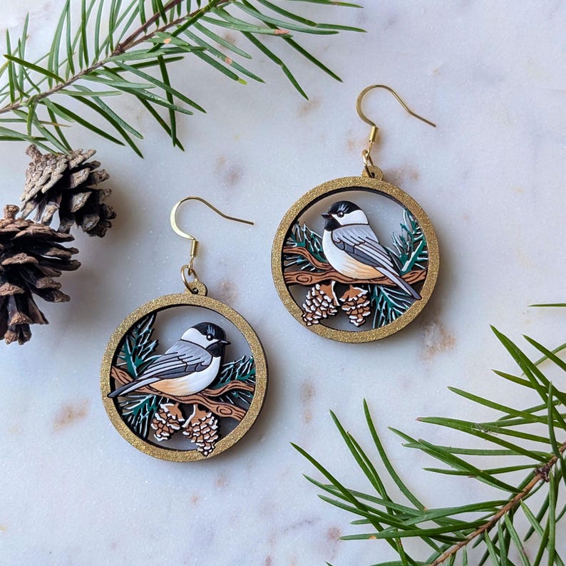 Hand-painted, laser-etched winter chickadee earrings which are 2 layers of wood with a round golden frame. The frame holds a scene with snow-capped pine needles and a chickadee perched on a branch next to pine cones.