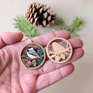 May include: Two circular wooden dangle earrings featuring a chickadee bird perched on a branch with pine needles and cones. One earring is painted in natural colors, while the other shows the unpainted laser-cut wood design.