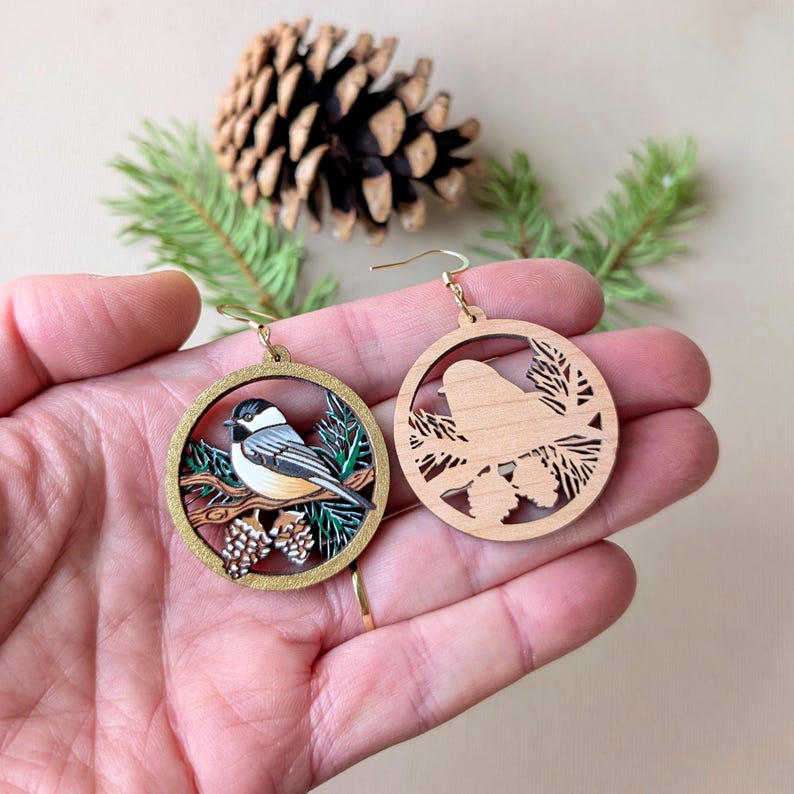 May include: Two circular wooden dangle earrings featuring a chickadee bird perched on a branch with pine needles and cones. One earring is painted in natural colors, while the other shows the unpainted laser-cut wood design.