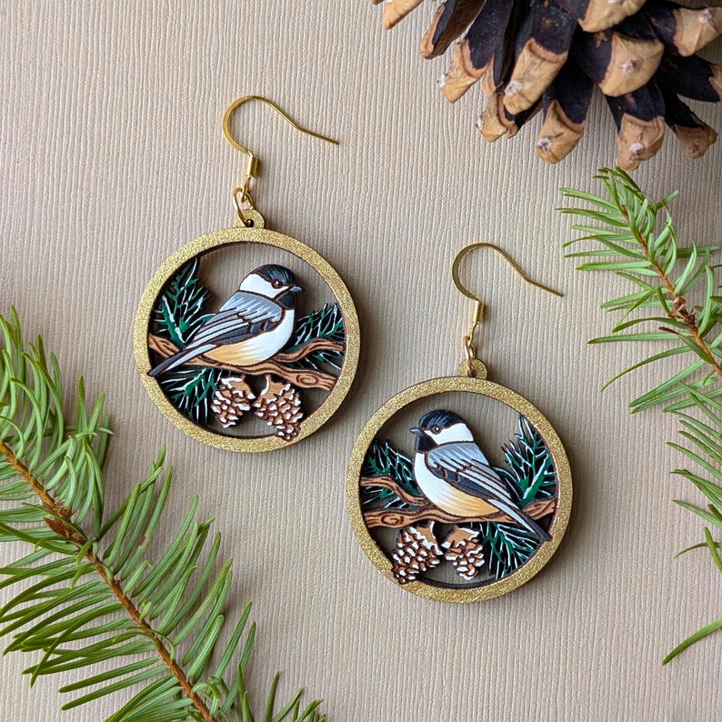 Hand-painted, laser-etched winter chickadee earrings which are 2 layers of wood with a round golden frame. The frame holds a scene with snow-capped pine needles and a chickadee perched on a branch next to pine cones.