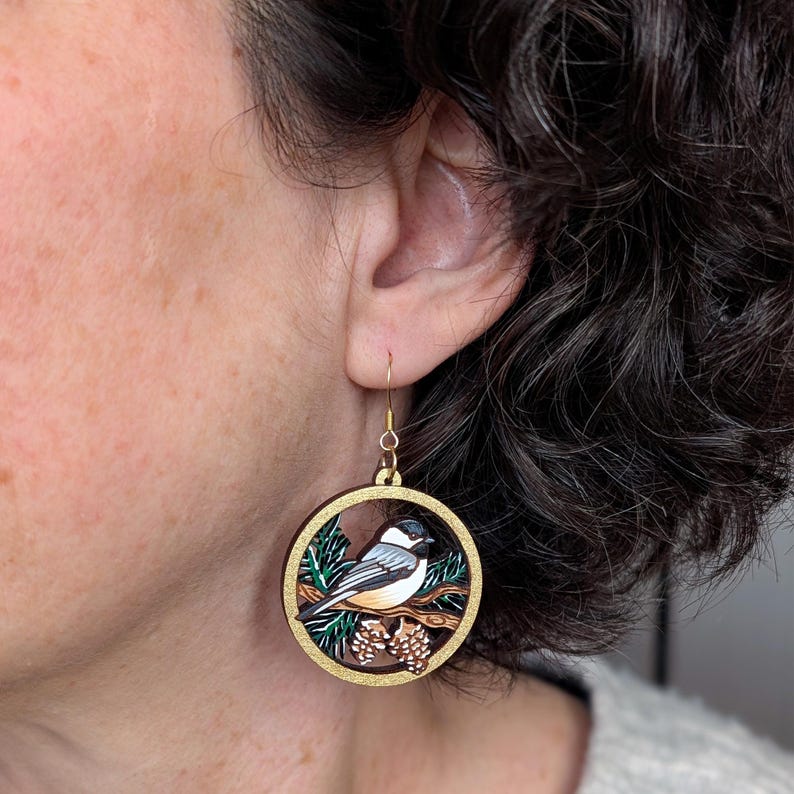 May include: A circular wooden dangle earring featuring a detailed illustration of a black-capped chickadee perched on a pine branch with pinecones. The design is framed by a gold-toned border and hangs from a simple gold-toned metal hook.