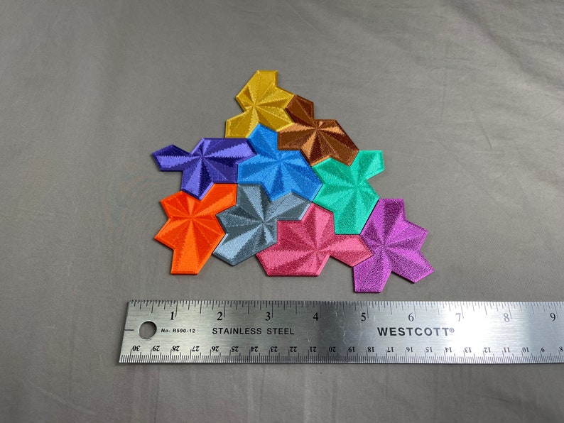 May include: A set of seven colorful geometric puzzle pieces, each with a starburst design. The pieces are arranged in a triangular shape, with the largest piece at the top and the smallest piece at the bottom. The pieces are made of a textured material and have a metallic sheen.