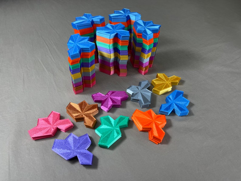 May include: A collection of colorful, star-shaped tiles made of plastic. The tiles are stacked in piles of different heights, with some piles arranged in a row. The tiles are in various colors, including blue, green, purple, orange, and gold.