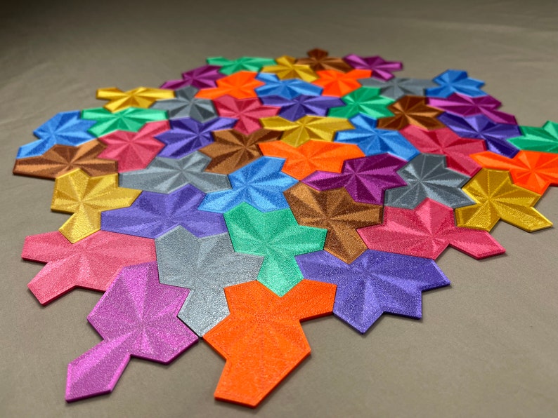 May include: A close-up of a colorful geometric puzzle made of 3D printed tiles. The tiles are arranged in a pattern and have a shiny, metallic finish. The colors include gold, silver, purple, pink, orange, blue, green, and brown.