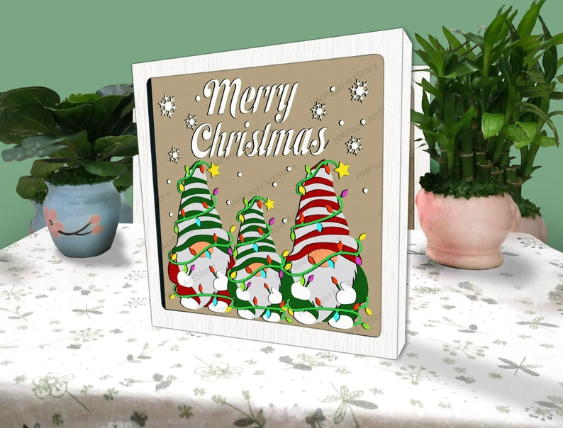 以下が含まれることがあります： 「メリークリスマス」と書かれた木製の看板と、赤と緑の帽子をかぶってクリスマスツリーを持った光るライトを持った3人のノームの楽しいシーン。ノームは雪片のある茶色の背景に立っています。