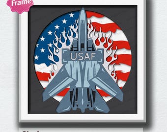 May include: A multi-layered shadow box art piece featuring a blue and grey fighter jet with the text USAF on its fuselage. The jet is centered over a circular background depicting the American flag with white flame-like cutouts surrounding it.